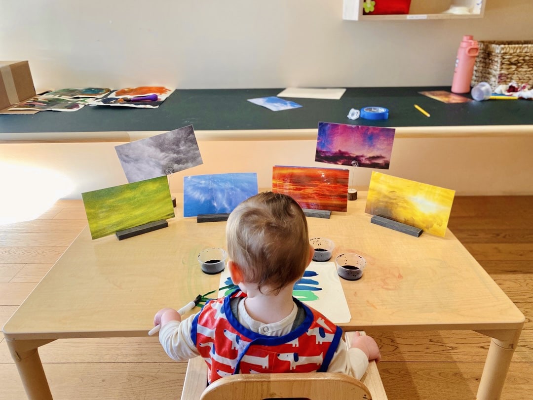 Young child painting at a table, surrounded by vibrant nature scenes, exploring creative expression through art.